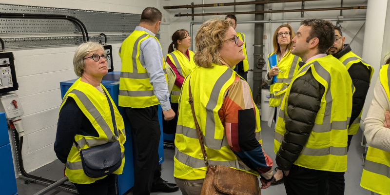 A group of people wearing yellow vests stands together in a medical gas plant room, engaged in conversation or discussion.