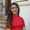 A portrait of Dhanika Kapoor stood next a white stone building, she is wearing a red dress.