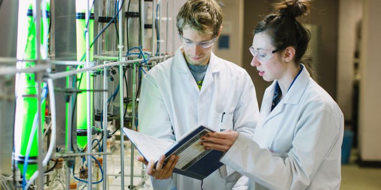Two students wearing white coats in a lab, reading notes from a folder while standing next to glass tubes filled with green liquid
