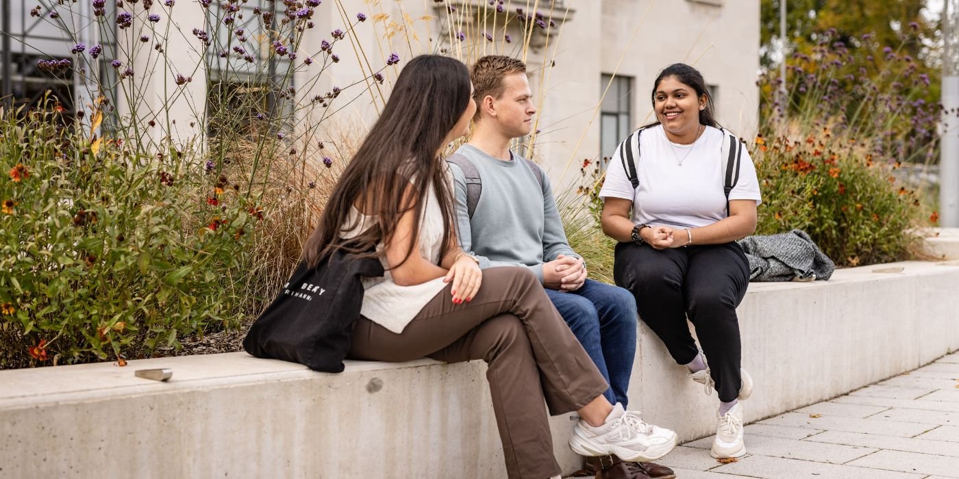 Three students sat on a wall in the garden, with the Bragg building behind them.