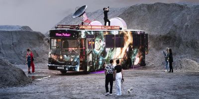 The Moon Palace bus observatory on a moon-like surface surrounded by people, with a child on top looking into the skies through binoculars