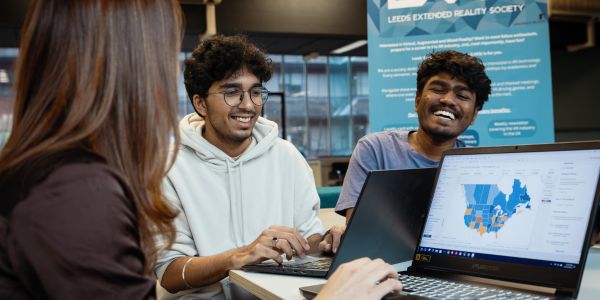 Group of 3 students laughing around a table with a laptop showing a graph
