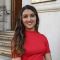 A portrait of Dhanika Kapoor stood next a white stone building, she is wearing a red dress.