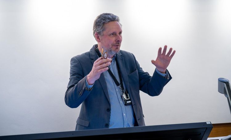 Professor Rob Dwyer-Joyce, with his arms raised, speaking to an unseen audience from behind a desk.